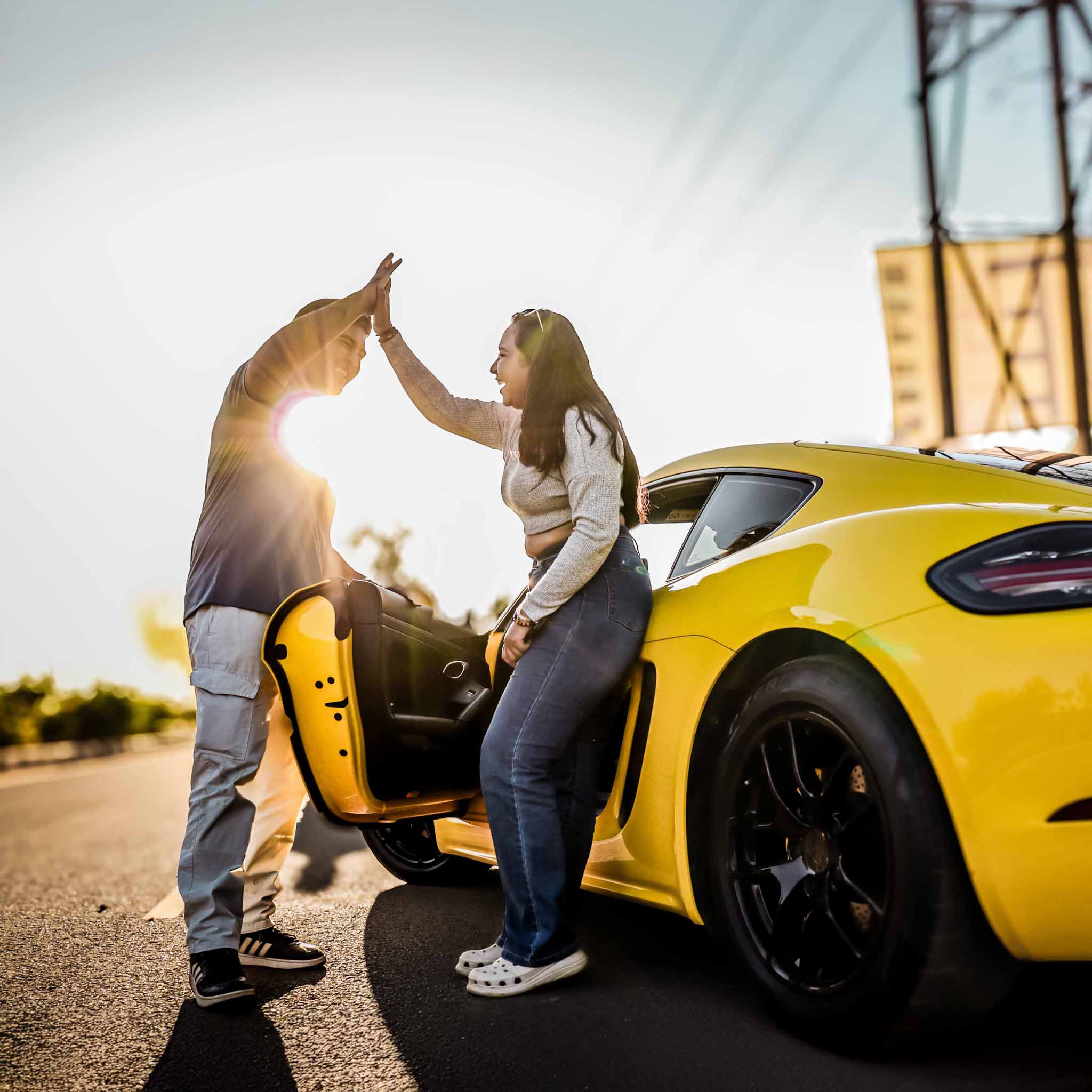 Two people celebrating with high five next to a yellow Porsche 718 Cayman at sunset